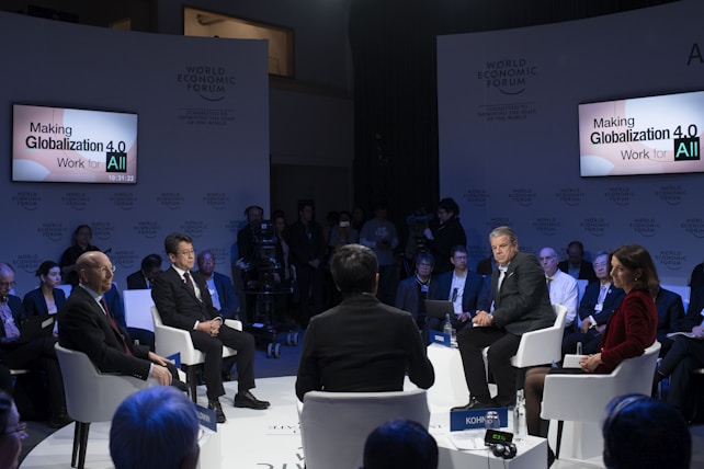 A formal panel discussion taking place at the World Economic Forum. Several individuals are seated in a circle, engaging in conversation while a small audience observes. Two screens display the text 'Making Globalization 4.0 Work for All'. The participants appear to be business professionals or academics.