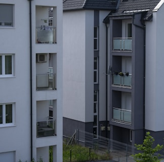 Several modern apartment buildings with balconies featuring glass railings. The structures have a clean and minimalist design with grey and white color tones. A small patch of grass is visible at the base.