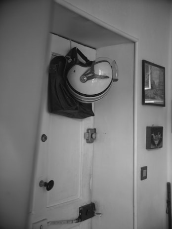 A motorcycle helmet with a visor is hung on a hook over a black bag, which appears to be attached to the top of a white, wooden door. The door is equipped with a door knob, a lock, and a latch. There are two framed pictures on the wall to the right of the door.