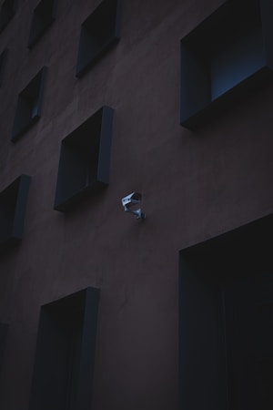 A dark, monochromatic building wall with recessed square windows. A security camera is mounted on the wall, adding a sense of surveillance and modern architecture.