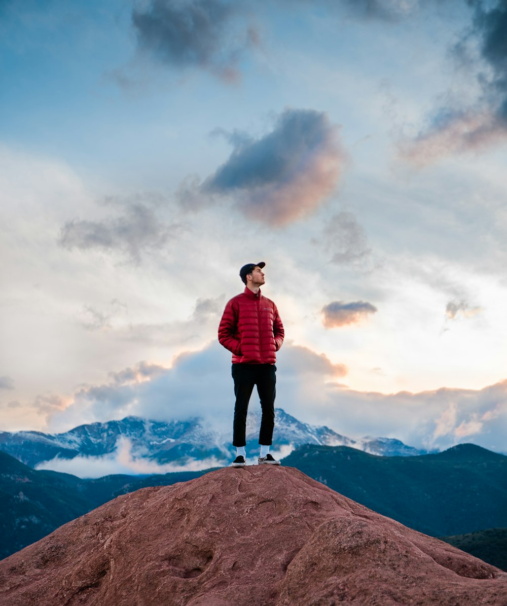 man in red jacket, black pants and black fitted cap standing on top of brown hill