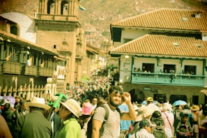 A bustling street crowded with people in traditional attire, set against the backdrop of historic architecture featuring terracotta rooftops and stone structures. A mix of tourists and locals navigate through the vibrant marketplace atmosphere.