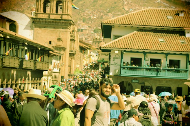 A bustling street crowded with people in traditional attire, set against the backdrop of historic architecture featuring terracotta rooftops and stone structures. A mix of tourists and locals navigate through the vibrant marketplace atmosphere.