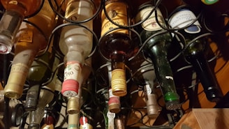 A collection of wine bottles stored horizontally in a metal wine rack. The arrangement displays various labels and colors of the bottles, indicating a diverse selection of wines. The bottles vary in color from clear to dark green and brown.