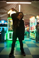 A silhouetted figure wearing a hockey mask with glowing red eyes stands ominously in a dimly lit arcade. Brightly lit arcade machines surround the figure, casting colorful neon lights and creating an eerie atmosphere. People are seen in the background, adding a sense of liveliness to the scene.