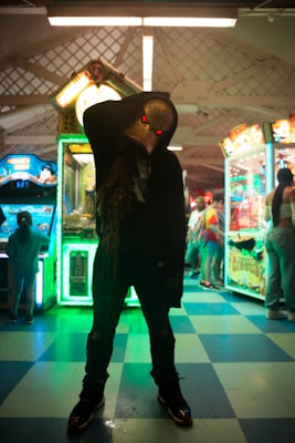 A silhouetted figure wearing a hockey mask with glowing red eyes stands ominously in a dimly lit arcade. Brightly lit arcade machines surround the figure, casting colorful neon lights and creating an eerie atmosphere. People are seen in the background, adding a sense of liveliness to the scene.
