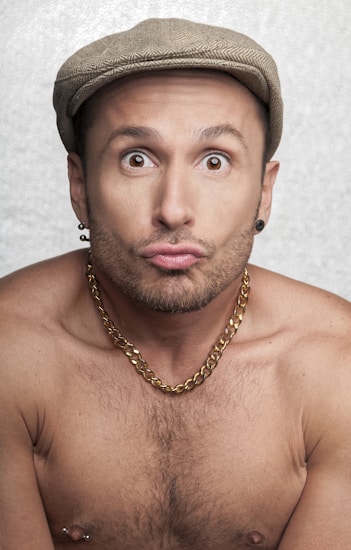 A shirtless person is wearing a beige cap and a thick gold chain necklace, making a playful face with wide eyes and puckered lips. The background is a light, textured surface.