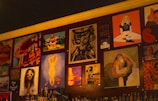 A wall adorned with various pieces of artwork, showcasing diverse styles and subjects. The artwork includes abstract designs, portraits, and graphic typography. Below the artworks, bottles are arranged, suggesting it might be a bar or café setting.