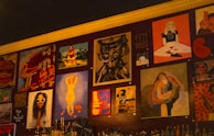 A wall adorned with various pieces of artwork, showcasing diverse styles and subjects. The artwork includes abstract designs, portraits, and graphic typography. Below the artworks, bottles are arranged, suggesting it might be a bar or café setting.