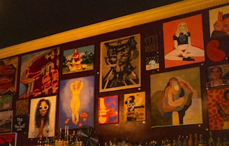 A wall adorned with various pieces of artwork, showcasing diverse styles and subjects. The artwork includes abstract designs, portraits, and graphic typography. Below the artworks, bottles are arranged, suggesting it might be a bar or café setting.
