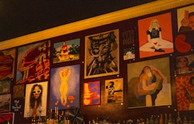 A wall adorned with various pieces of artwork, showcasing diverse styles and subjects. The artwork includes abstract designs, portraits, and graphic typography. Below the artworks, bottles are arranged, suggesting it might be a bar or café setting.