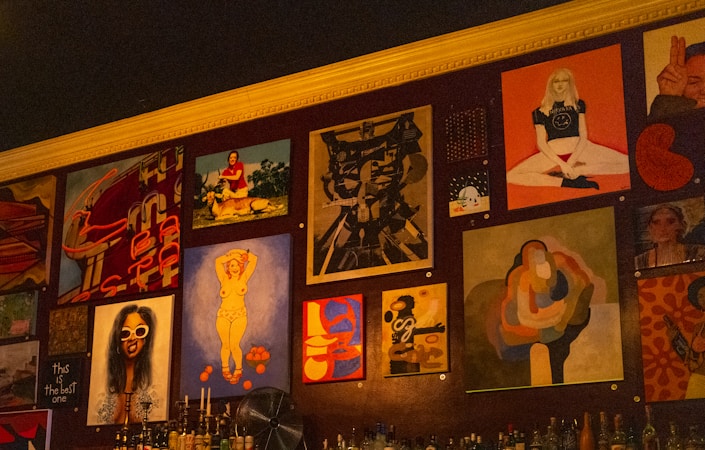 A wall adorned with various pieces of artwork, showcasing diverse styles and subjects. The artwork includes abstract designs, portraits, and graphic typography. Below the artworks, bottles are arranged, suggesting it might be a bar or café setting.