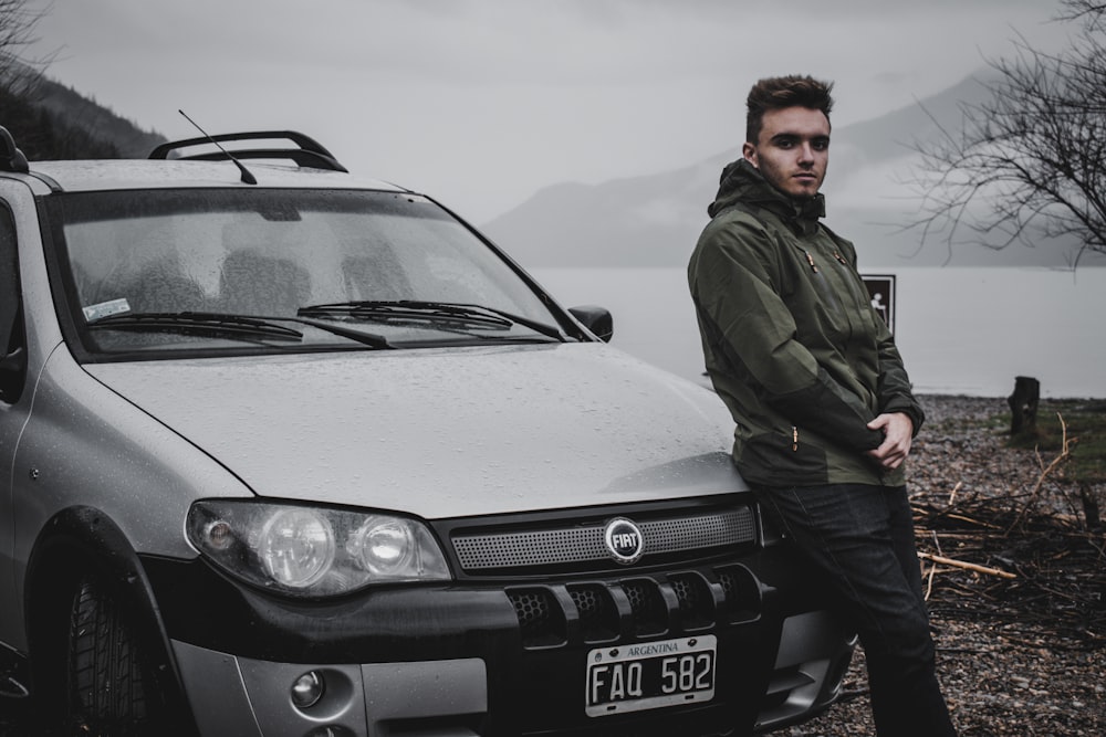 man in green jacket and black pants leaning on parked silver Fiat car