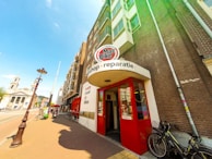 A street scene featuring a brick building with a bike shop entrance. The shop's signage includes 'Mac Bike' and 'reparatie' above the red entrance doors. Bicycles are parked outside on the sidewalk. The street also features a traditional lamp post, with a clear blue sky overhead and a church visible in the background.