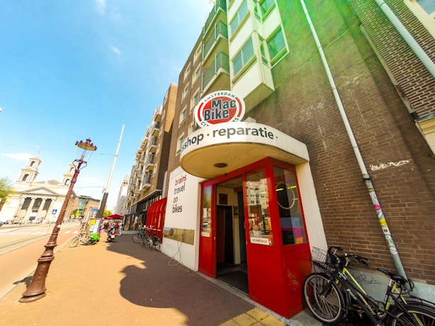 A street scene featuring a brick building with a bike shop entrance. The shop's signage includes 'Mac Bike' and 'reparatie' above the red entrance doors. Bicycles are parked outside on the sidewalk. The street also features a traditional lamp post, with a clear blue sky overhead and a church visible in the background.