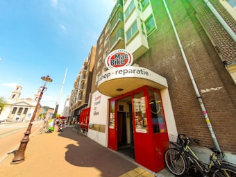 A street scene featuring a brick building with a bike shop entrance. The shop's signage includes 'Mac Bike' and 'reparatie' above the red entrance doors. Bicycles are parked outside on the sidewalk. The street also features a traditional lamp post, with a clear blue sky overhead and a church visible in the background.