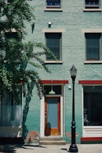 A green brick building with three windows on the upper level and a door on the ground level. The door is framed with white and has a glass panel, above which hangs a green light fixture. To the right of the door is a black street lamp. To the left, a tree with abundant green foliage partially covers the building. Red-brown trims are featured above the windows and door. Some areas on the lower part of the building show signs of peeling paint and exposed bricks.