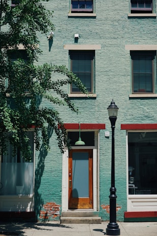 A green brick building with three windows on the upper level and a door on the ground level. The door is framed with white and has a glass panel, above which hangs a green light fixture. To the right of the door is a black street lamp. To the left, a tree with abundant green foliage partially covers the building. Red-brown trims are featured above the windows and door. Some areas on the lower part of the building show signs of peeling paint and exposed bricks.