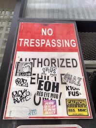 A 'No Trespassing' sign is prominently displayed, partially covered with stickers showing various designs, logos, and texts. The red and white sign is affixed to a surface, possibly near a building or entrance.