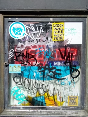 A framed glass surface covered with a variety of colorful graffiti and stickers. Bright red LED numbers are partly visible behind the writing. The stickers display text and abstract designs, and the graffiti is layered, with different styles and colors overlapping each other.