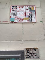 A wall featuring a large sign covered with a variety of graffiti stickers and tags. The stickers have diverse styles, including logos and hand-drawn elements. Below this, a smaller sign exhibits more graffiti writing in black paint, creating an urban and gritty aesthetic.