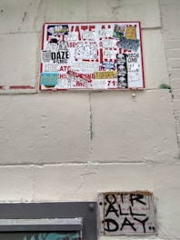 A wall featuring a large sign covered with a variety of graffiti stickers and tags. The stickers have diverse styles, including logos and hand-drawn elements. Below this, a smaller sign exhibits more graffiti writing in black paint, creating an urban and gritty aesthetic.
