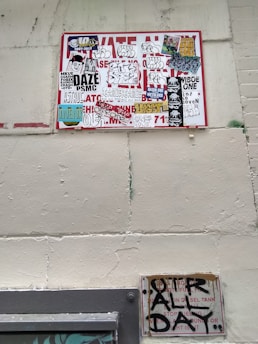 A wall featuring a large sign covered with a variety of graffiti stickers and tags. The stickers have diverse styles, including logos and hand-drawn elements. Below this, a smaller sign exhibits more graffiti writing in black paint, creating an urban and gritty aesthetic.