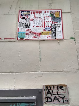 A wall featuring a large sign covered with a variety of graffiti stickers and tags. The stickers have diverse styles, including logos and hand-drawn elements. Below this, a smaller sign exhibits more graffiti writing in black paint, creating an urban and gritty aesthetic.