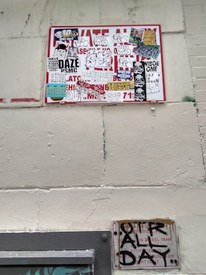 A wall featuring a large sign covered with a variety of graffiti stickers and tags. The stickers have diverse styles, including logos and hand-drawn elements. Below this, a smaller sign exhibits more graffiti writing in black paint, creating an urban and gritty aesthetic.