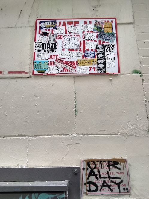 A wall featuring a large sign covered with a variety of graffiti stickers and tags. The stickers have diverse styles, including logos and hand-drawn elements. Below this, a smaller sign exhibits more graffiti writing in black paint, creating an urban and gritty aesthetic.