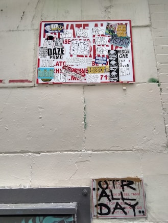 A wall featuring a large sign covered with a variety of graffiti stickers and tags. The stickers have diverse styles, including logos and hand-drawn elements. Below this, a smaller sign exhibits more graffiti writing in black paint, creating an urban and gritty aesthetic.