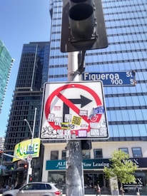 A no right turn traffic sign covered with various colorful stickers is mounted on a metallic pole. Tall glass skyscrapers and a street sign for Figueroa Street are visible in the background under a clear blue sky. The base of the signpost is weathered, and a traffic light is attached to it. Cars and urban buildings with signage are present at street level.