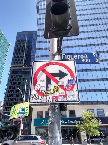 A no right turn traffic sign covered with various colorful stickers is mounted on a metallic pole. Tall glass skyscrapers and a street sign for Figueroa Street are visible in the background under a clear blue sky. The base of the signpost is weathered, and a traffic light is attached to it. Cars and urban buildings with signage are present at street level.
