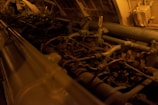 A complex array of machinery parts, including pipes, tubes, and valves, illuminated in a dim, warm light. The setting appears industrial, with metallic and mechanical components arranged within a confined space.