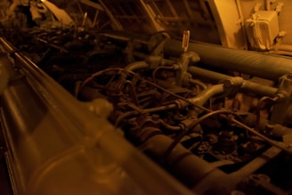 A complex array of machinery parts, including pipes, tubes, and valves, illuminated in a dim, warm light. The setting appears industrial, with metallic and mechanical components arranged within a confined space.