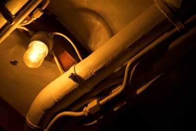 A warm light bulb illuminates a series of intertwined pipes and cables, casting dramatic shadows on the ceiling. The scene has an industrial feel, with the lighting adding a cozy yet mysterious atmosphere.