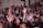 A large group of women appears to be engaged in a communal activity, with many raising their arms. Some are wearing floral crowns and there is a sense of togetherness and focus.