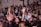 A large group of women appears to be engaged in a communal activity, with many raising their arms. Some are wearing floral crowns and there is a sense of togetherness and focus.