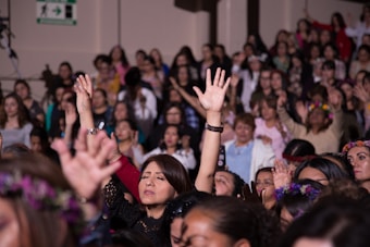 A large group of women appears to be engaged in a communal activity, with many raising their arms. Some are wearing floral crowns and there is a sense of togetherness and focus.