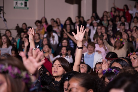 A large group of women appears to be engaged in a communal activity, with many raising their arms. Some are wearing floral crowns and there is a sense of togetherness and focus.