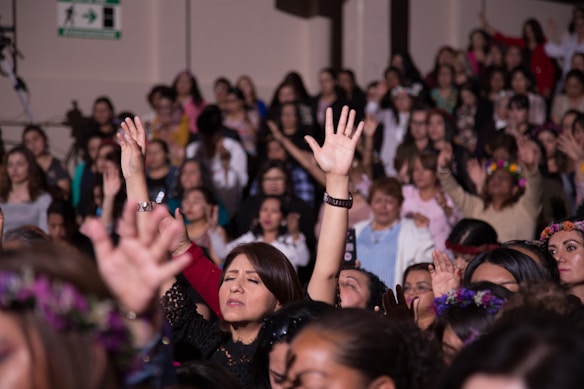A large group of women appears to be engaged in a communal activity, with many raising their arms. Some are wearing floral crowns and there is a sense of togetherness and focus.