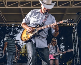 A guitarist wearing a cowboy hat energetically plays an electric guitar on stage, surrounded by other band members. Behind him, a woman is playing a pink bass guitar and a drummer is partially visible. The stage is set outdoors with lighting structures and a backdrop of trees.