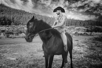 A person wearing a cowboy hat is riding a dark horse in a natural landscape. The background features a dense forest and a cloudy sky. The image is in black and white, creating a timeless and dramatic effect.