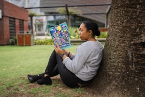 A person sitting on the grass, leaning against a tree while holding a colorful magazine. The background consists of a garden area with modern buildings and greenery.