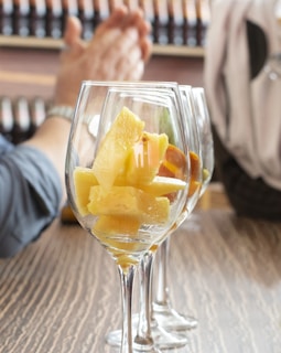 Several wine glasses are aligned on a wooden table, each filled with chunks of yellow fruit, possibly pineapple or similar. In the background, a blurred figure appears to be clapping or gesturing with hands together. Bottles are arranged on a shelf, slightly out of focus, suggesting a setting like a bar or winery.