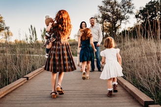 A family of five walks along a wooden pathway in a scenic, natural setting. The parents stand facing the camera, holding hands, while their three children, dressed in vintage-style clothing, walk ahead of them. The sun casts a warm glow over the grassy field around them and the late afternoon sky.