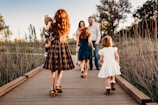 A family of five walks along a wooden pathway in a scenic, natural setting. The parents stand facing the camera, holding hands, while their three children, dressed in vintage-style clothing, walk ahead of them. The sun casts a warm glow over the grassy field around them and the late afternoon sky.