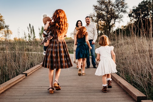 A family of five walks along a wooden pathway in a scenic, natural setting. The parents stand facing the camera, holding hands, while their three children, dressed in vintage-style clothing, walk ahead of them. The sun casts a warm glow over the grassy field around them and the late afternoon sky.