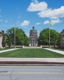 A symmetrical view of an academic building with a prominent dome, situated at the end of a well-maintained lawn lined with pathways. Two flagpoles, one with an American flag, flank the entrance. The landscape is complemented by lush green trees and a clear blue sky with a few clouds.
