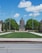 A symmetrical view of an academic building with a prominent dome, situated at the end of a well-maintained lawn lined with pathways. Two flagpoles, one with an American flag, flank the entrance. The landscape is complemented by lush green trees and a clear blue sky with a few clouds.
